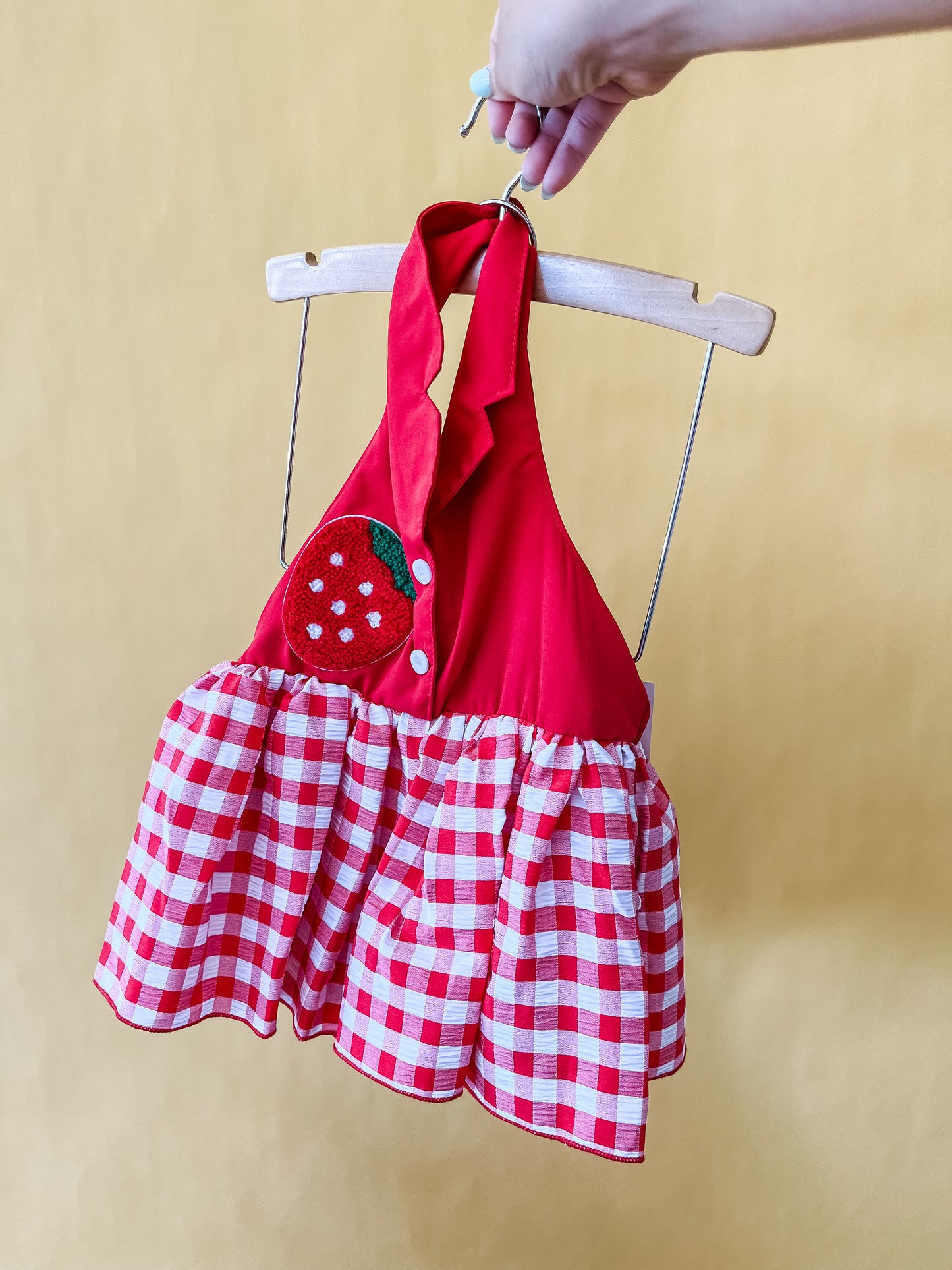 A hand holds up the Berry Darlin Romper by Tiny Tot Bundles, featuring a red and white checkered pattern with a halter neck and a strawberry patch on the chest, displayed on a wooden hanger against a yellow background.