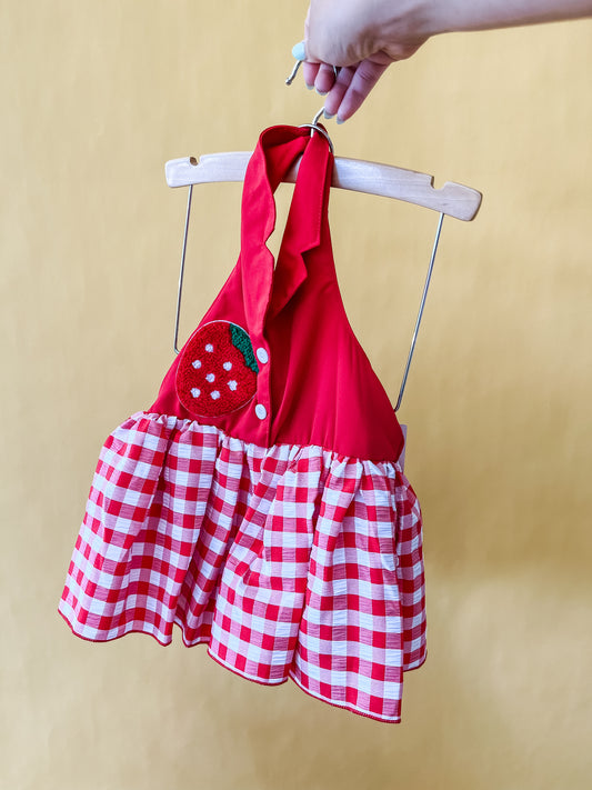A hand holds up the Berry Darlin Romper by Tiny Tot Bundles, featuring a red and white checkered pattern with a halter neck and a strawberry patch on the chest, displayed on a wooden hanger against a yellow background.