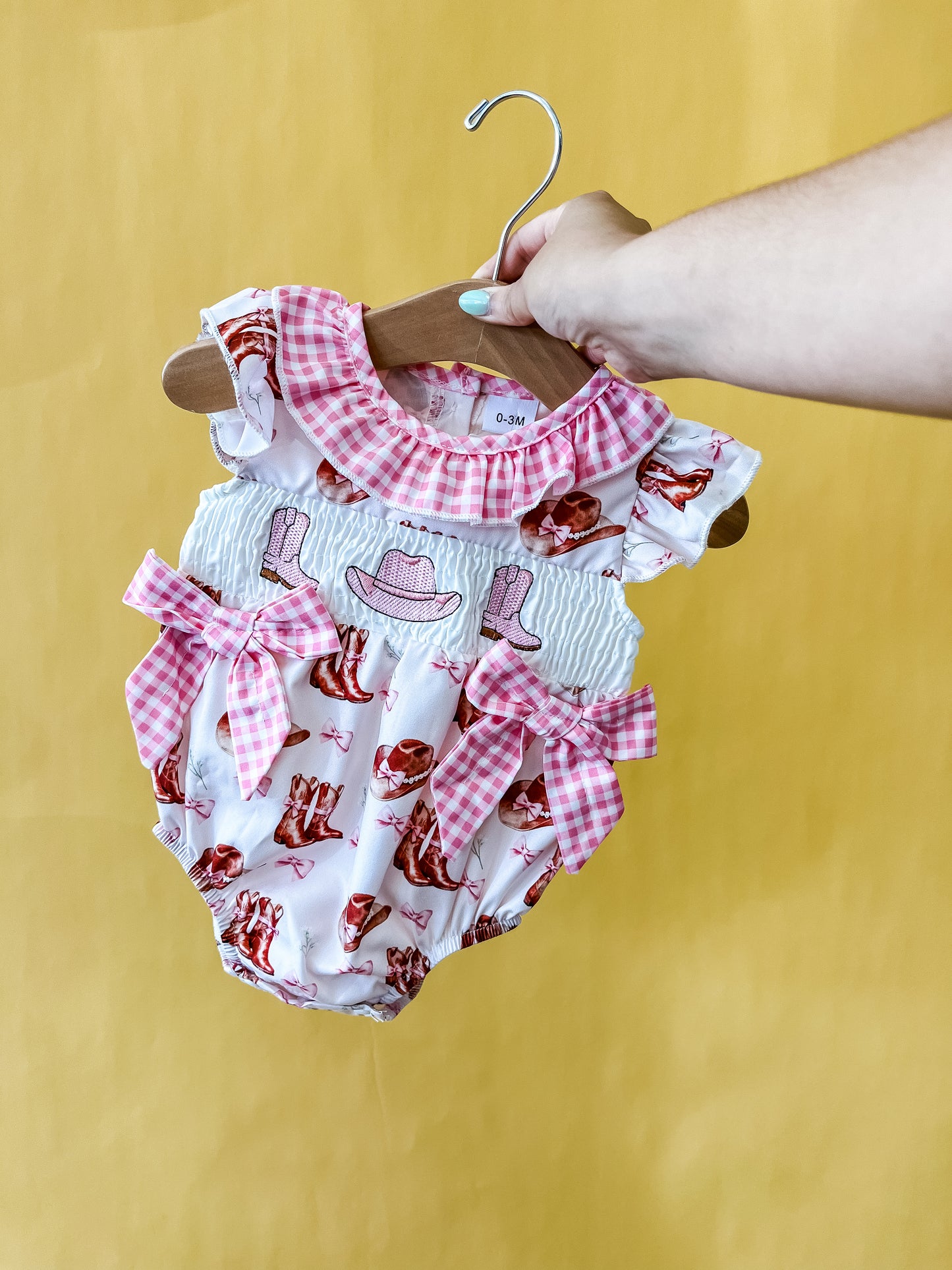 A hand holds the Tiny Tot Bundles Cowgirl Cutie Romper on a hanger. The white romper features pink gingham bows and collar, with cowgirl hats and boots printed on it, set against a yellow background.