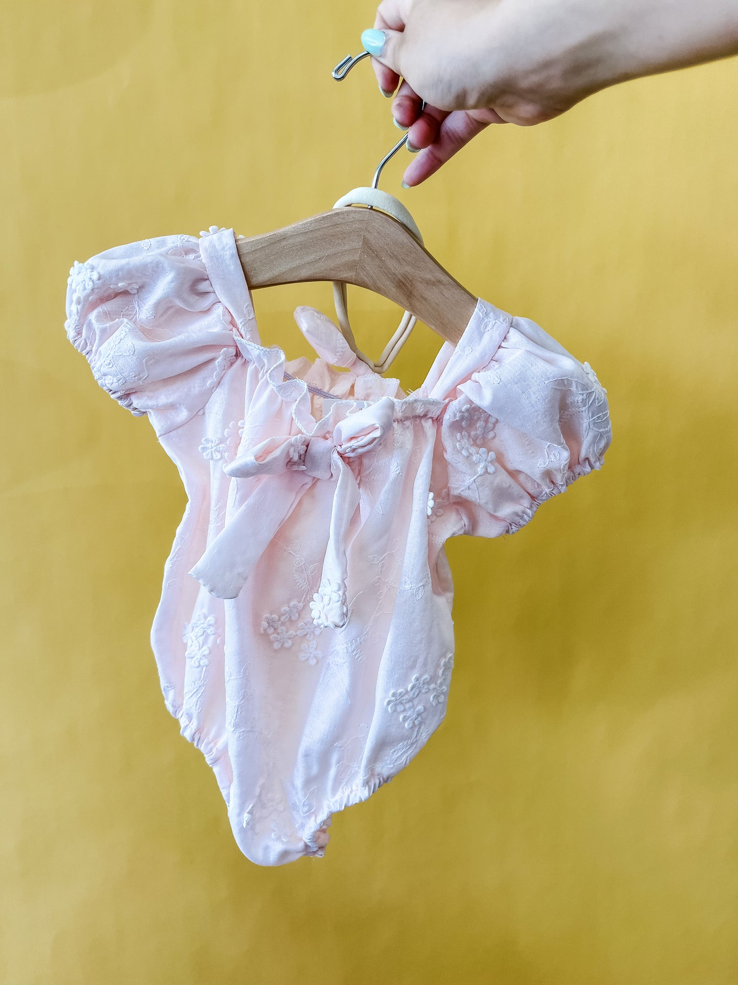 A hand holds a wooden hanger displaying the Tiny Tot Bundles Sweet Peony Bubble—a light pink baby onesie with puff sleeves, floral embroidery, and a bow—set against a yellow background.