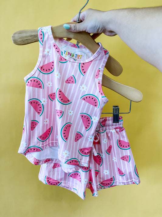 A hand holds a hanger displaying the Tiny Tot Bundles Melon Blossom Tank Set—an adorable pink toddler outfit adorned with watermelon slices and flowers—set against a yellow background.