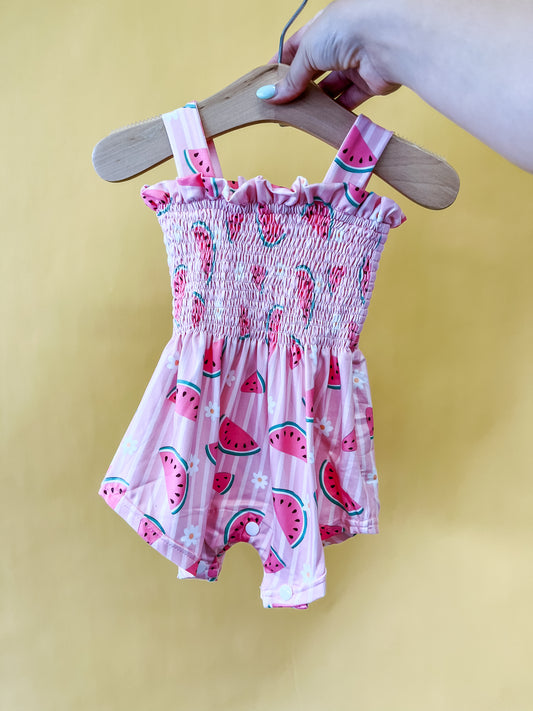 A hand holds a hanger with the Tiny Tot Bundles Melon Blossom Smocked Romper, featuring a watermelon print, against a solid yellow background.