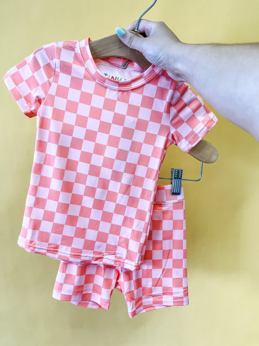 A hand holds a wooden hanger displaying the Coral Checkered Lounge Set from Tiny Tot Bundles, featuring a child's pink and white short-sleeve shirt with matching shorts, set against a yellow background.