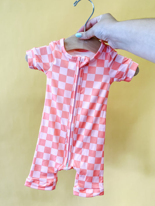 A hand holds the Tiny Tot Bundles Coral Checkered Short Zippy baby romper on a wooden hanger against a plain yellow background.
