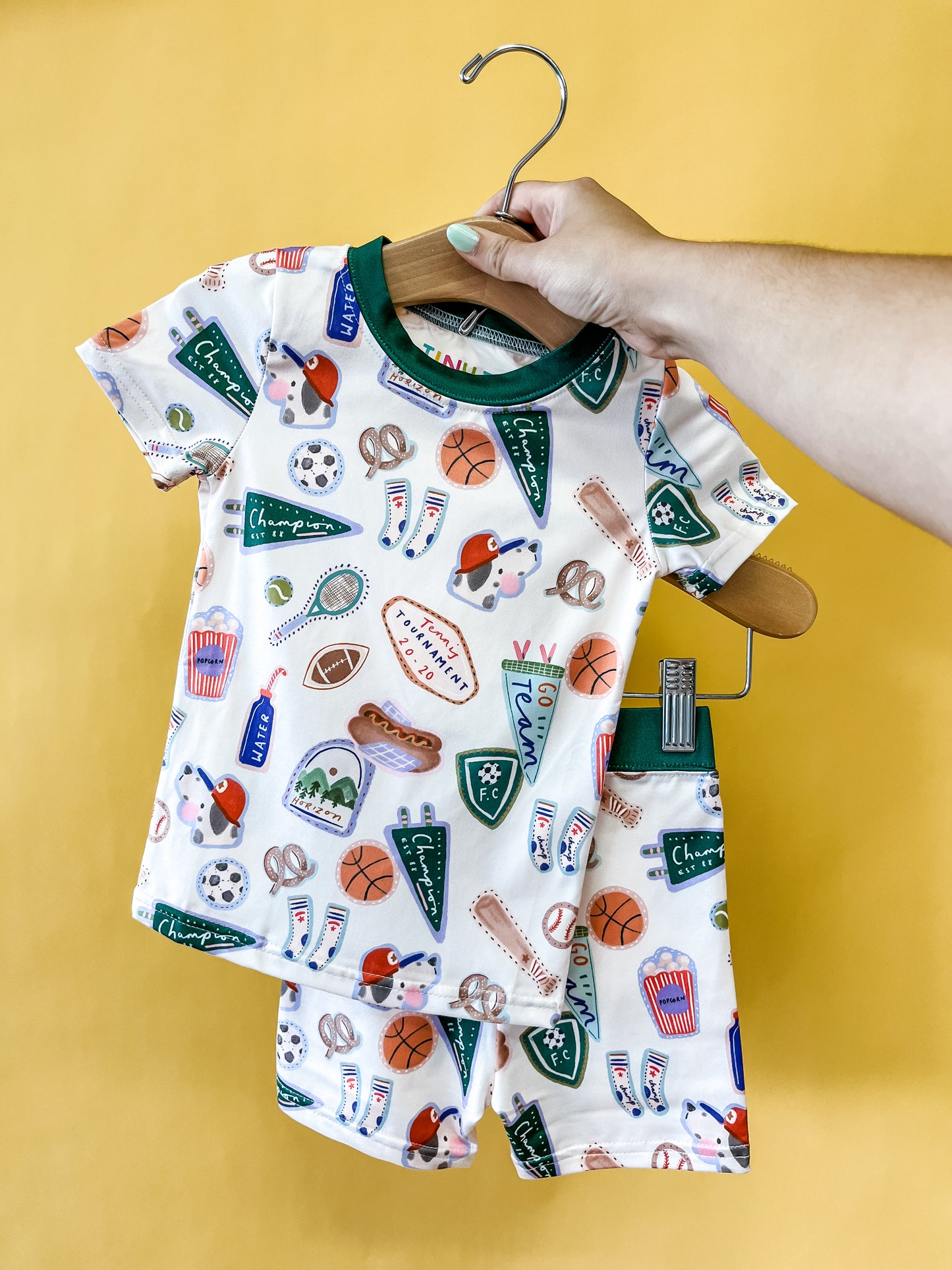 A hand holds the Tiny Tot Bundles All-Star Adventure Lounge Set for children, featuring pajamas decorated with colorful sports and stadium-themed illustrations—like balls, hats, popcorn, and pennants—set against a yellow background.