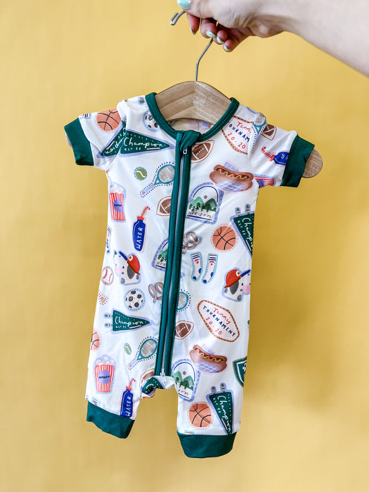 A hand holds a hanger with the Tiny Tot Bundles All-Star Adventure Short Zippy, a baby romper adorned with sports-themed prints including basketballs, hot dogs, and pennants, against a yellow background.