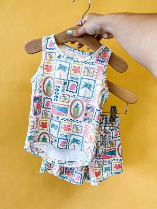 A hand holds a hanger displaying the Tiny Tot Bundles Sunshine Souvenir Tank Set—a colorful, tropical-print sleeveless toddler top with matching shorts—set against a bright yellow background.