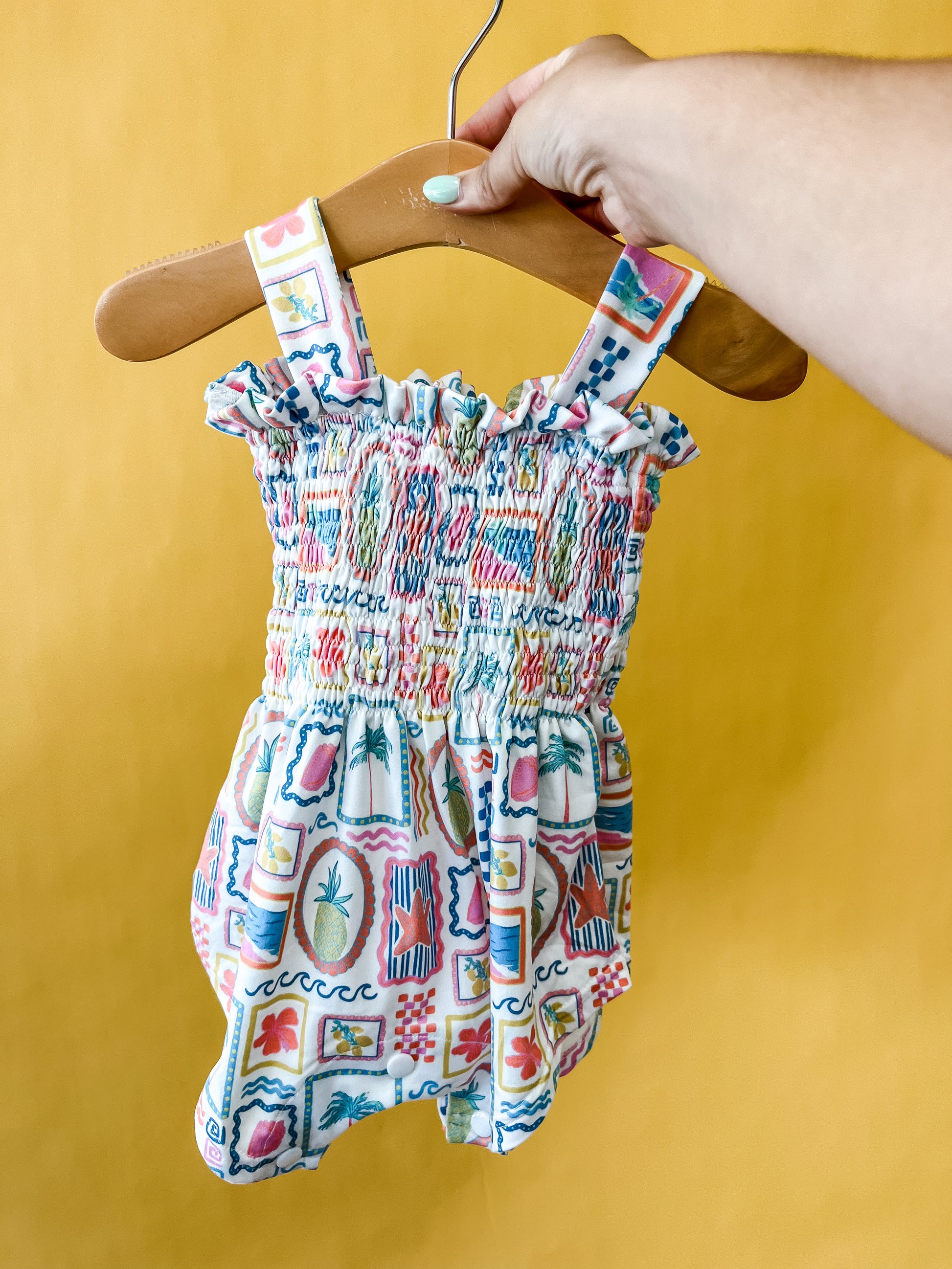 A hand holds a wooden hanger displaying the Tiny Tot Bundles Sunshine Souvenir Smocked Romper—a colorful, patterned baby romper with ruffled, smocked fabric and wide straps—against a bright yellow background.