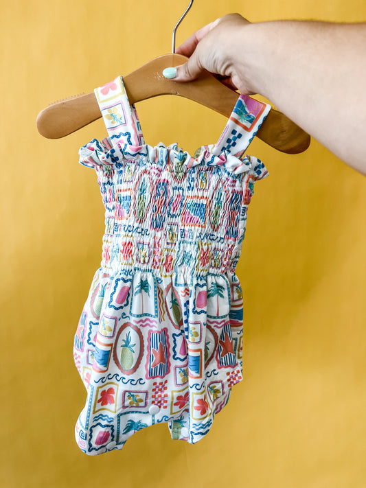 A hand holds a wooden hanger displaying the Tiny Tot Bundles Sunshine Souvenir Smocked Romper—a colorful, patterned baby romper with ruffled, smocked fabric and wide straps—against a bright yellow background.
