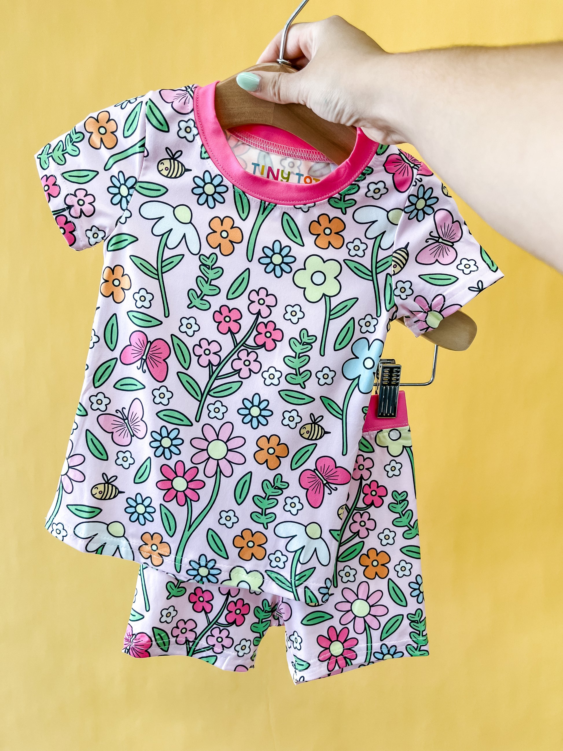 A hand holds up the Tiny Tot Bundles Honeybee Garden Lounge Set, featuring a baby outfit with a vibrant floral and butterfly pattern on a yellow background. The short-sleeve top and shorts are finished with pink trim around the neckline.