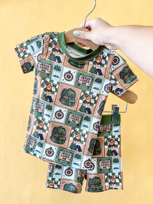 A hand holds the Tiny Tot Bundles Camper's Faith Lounge Set— a child’s matching t-shirt and shorts featuring camping-themed prints like tents, backpacks, pine trees, and “Jesus Loves Me” signs— against a yellow background.