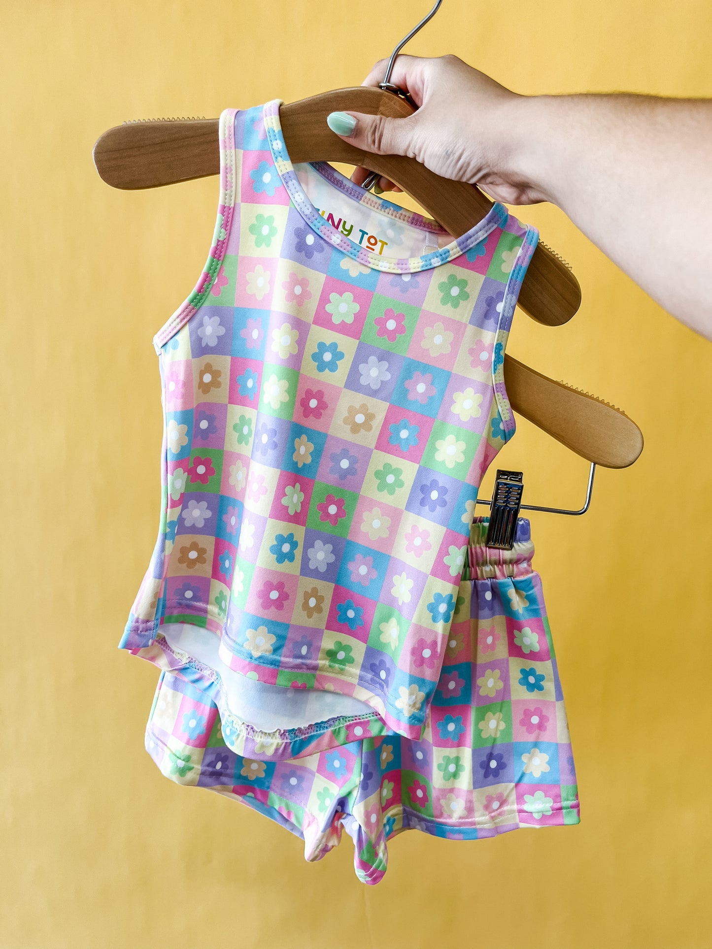 A hand holds a hanger with the Tiny Tot Bundles Pastel Petals Tank Set, featuring a pastel checkered tank top and shorts adorned with colorful flower patterns, against a yellow background.