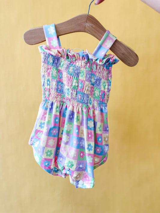 The Pastel Petals Smocked Romper by Tiny Tot Bundles, featuring charming floral and checkered patterns in pastel hues, hangs on a wooden hanger held by a hand against a yellow background.