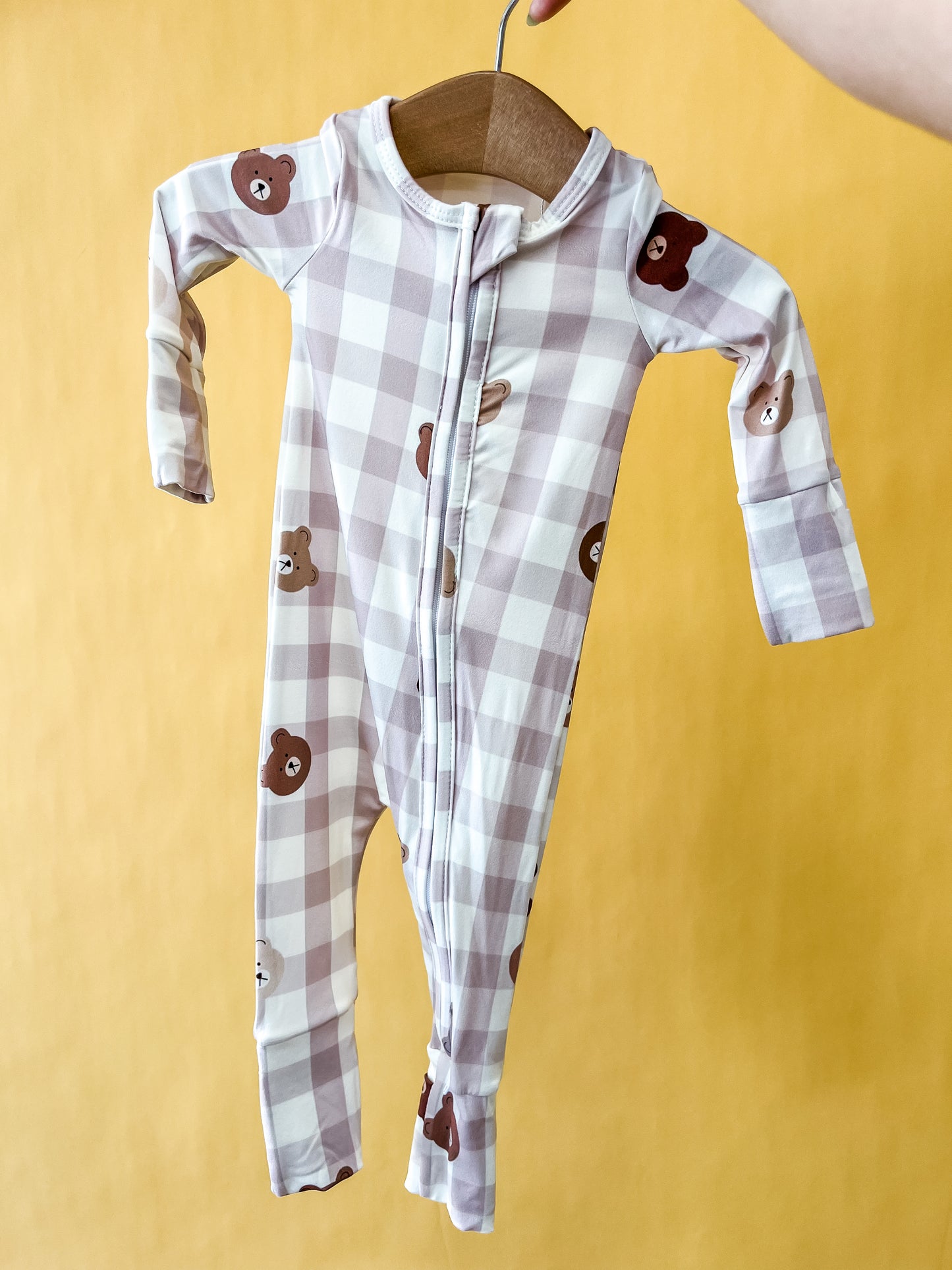 A hand holds a wooden hanger displaying the Tiny Tot Bundles Bear Picnic Long Zippy, a baby onesie adorned with a plaid pattern and brown teddy bear faces, set against a solid yellow background.