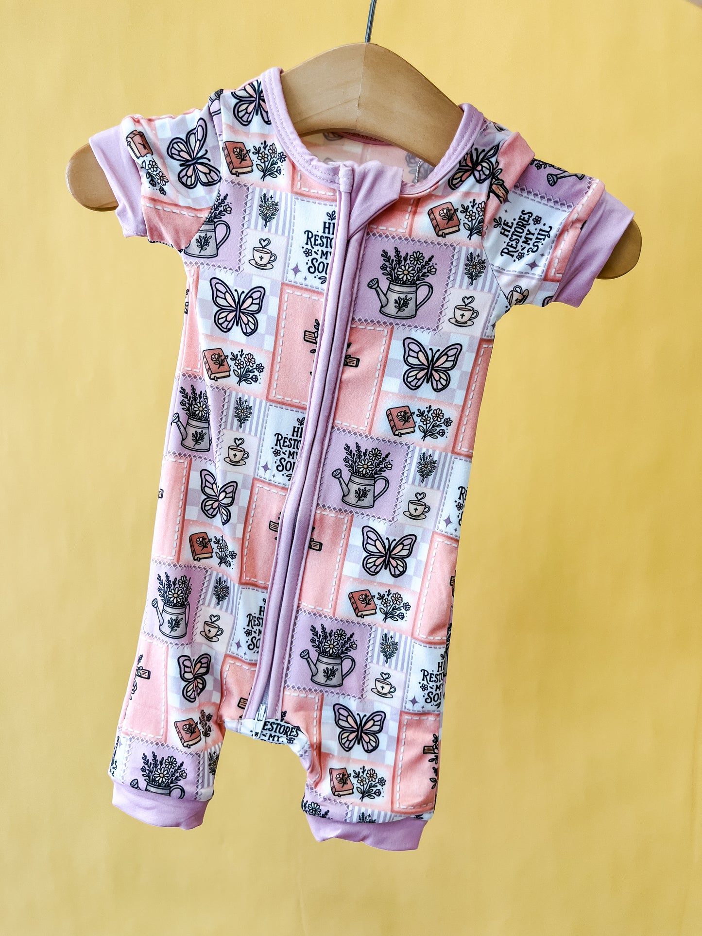 The Faith In Bloom Short Zippy by Tiny Tot Bundles, a short-sleeve baby romper featuring butterflies, teacups, and lavender plants on a pink and purple patchwork design, is displayed on a wooden hanger against a yellow background.