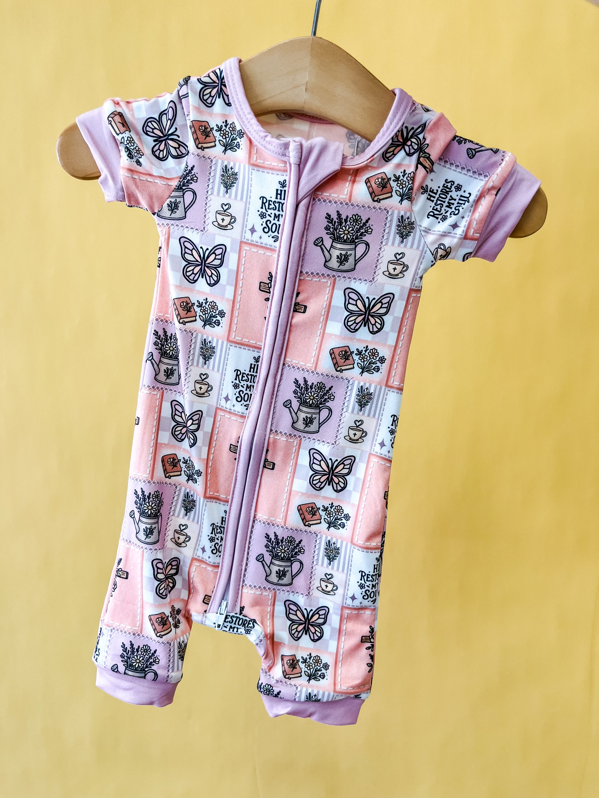 The Faith In Bloom Short Zippy by Tiny Tot Bundles, a short-sleeve baby romper featuring butterflies, teacups, and lavender plants on a pink and purple patchwork design, is displayed on a wooden hanger against a yellow background.