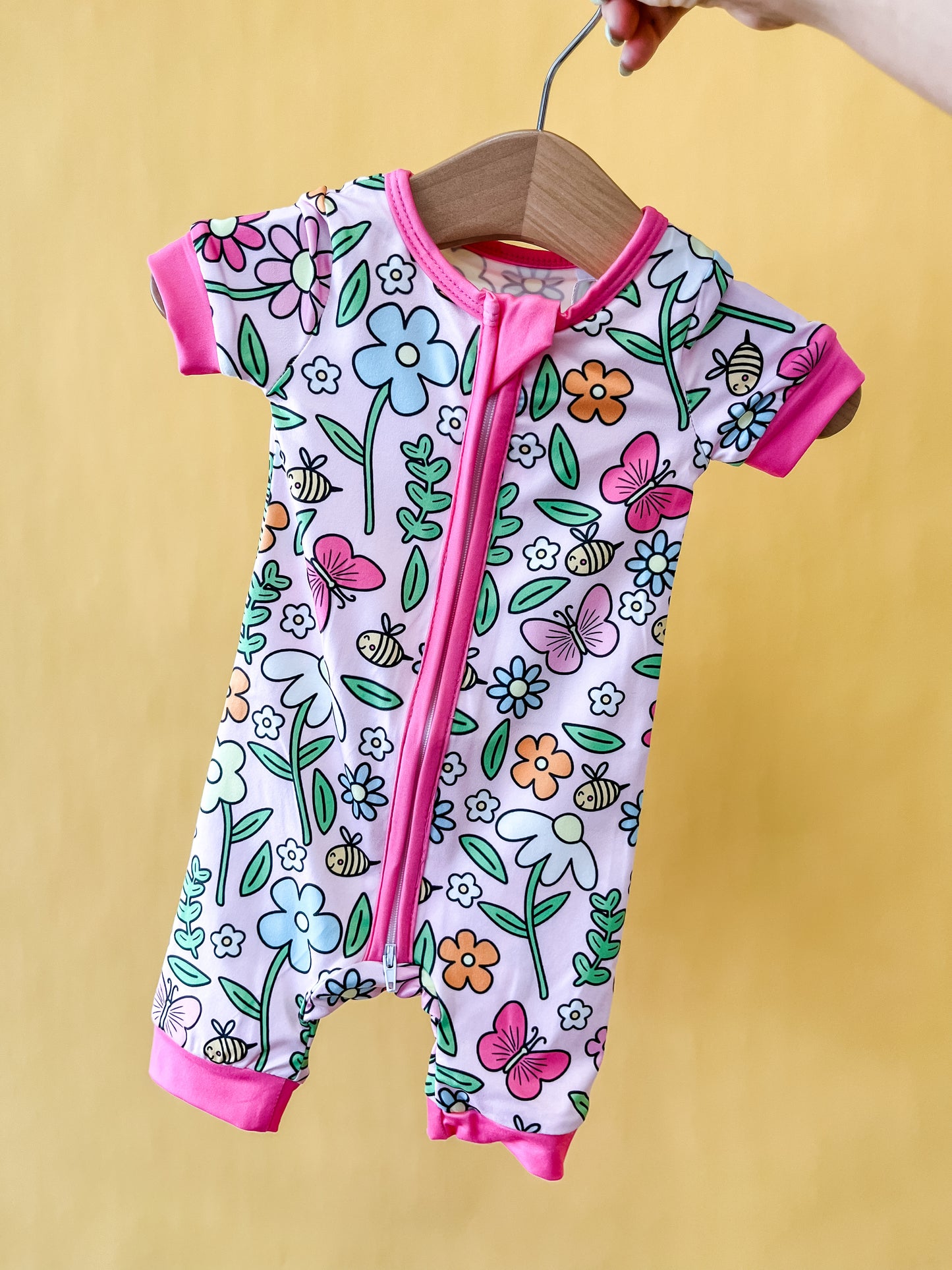 A hand holds the Honeybee Garden Short Zippy by Tiny Tot Bundles on a hanger. The romper features a colorful floral and butterfly pattern with pink trim and a front zipper, set against a yellow background.