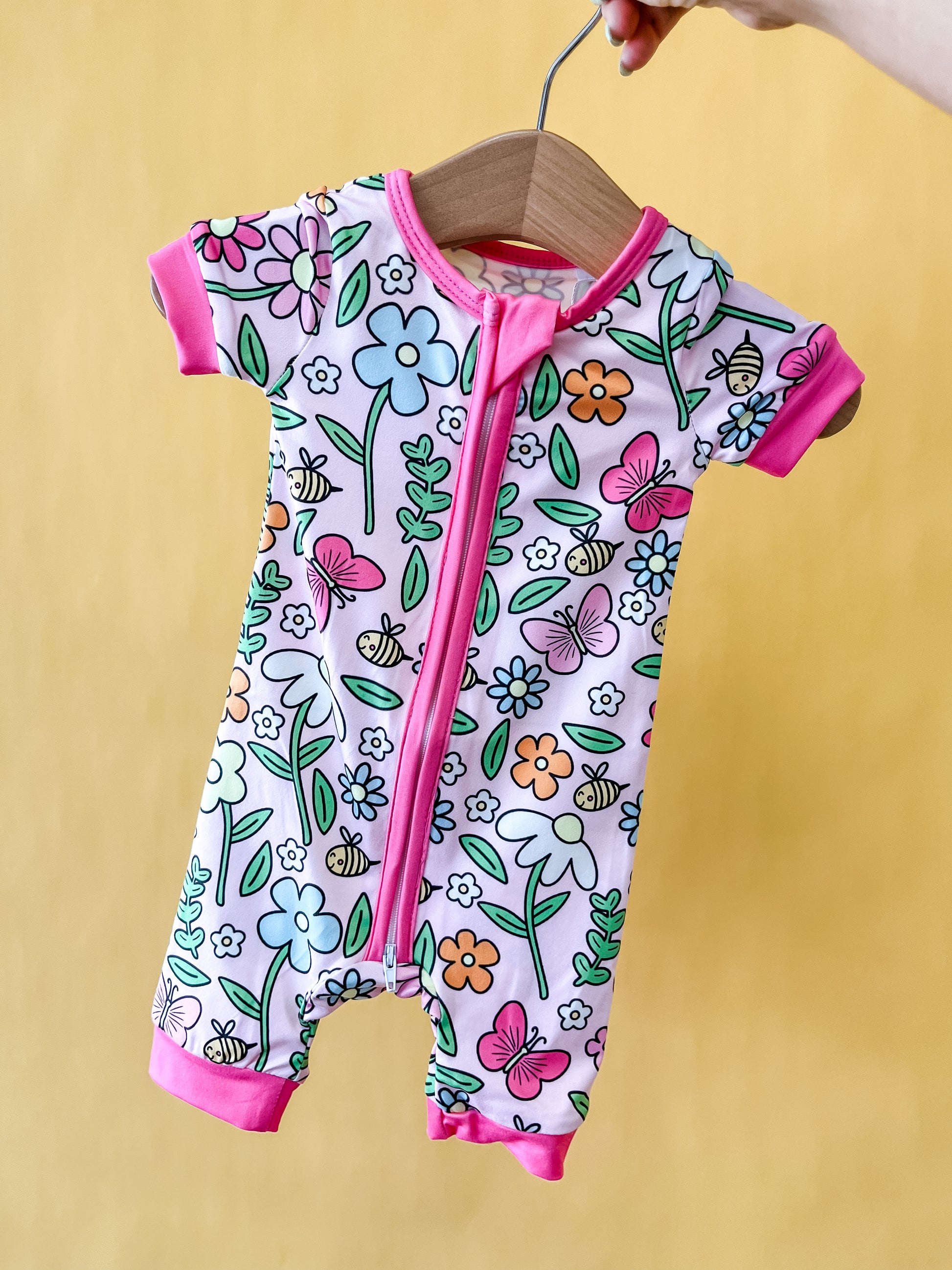 A hand holds the Honeybee Garden Short Zippy by Tiny Tot Bundles on a hanger. The romper features a colorful floral and butterfly pattern with pink trim and a front zipper, set against a yellow background.