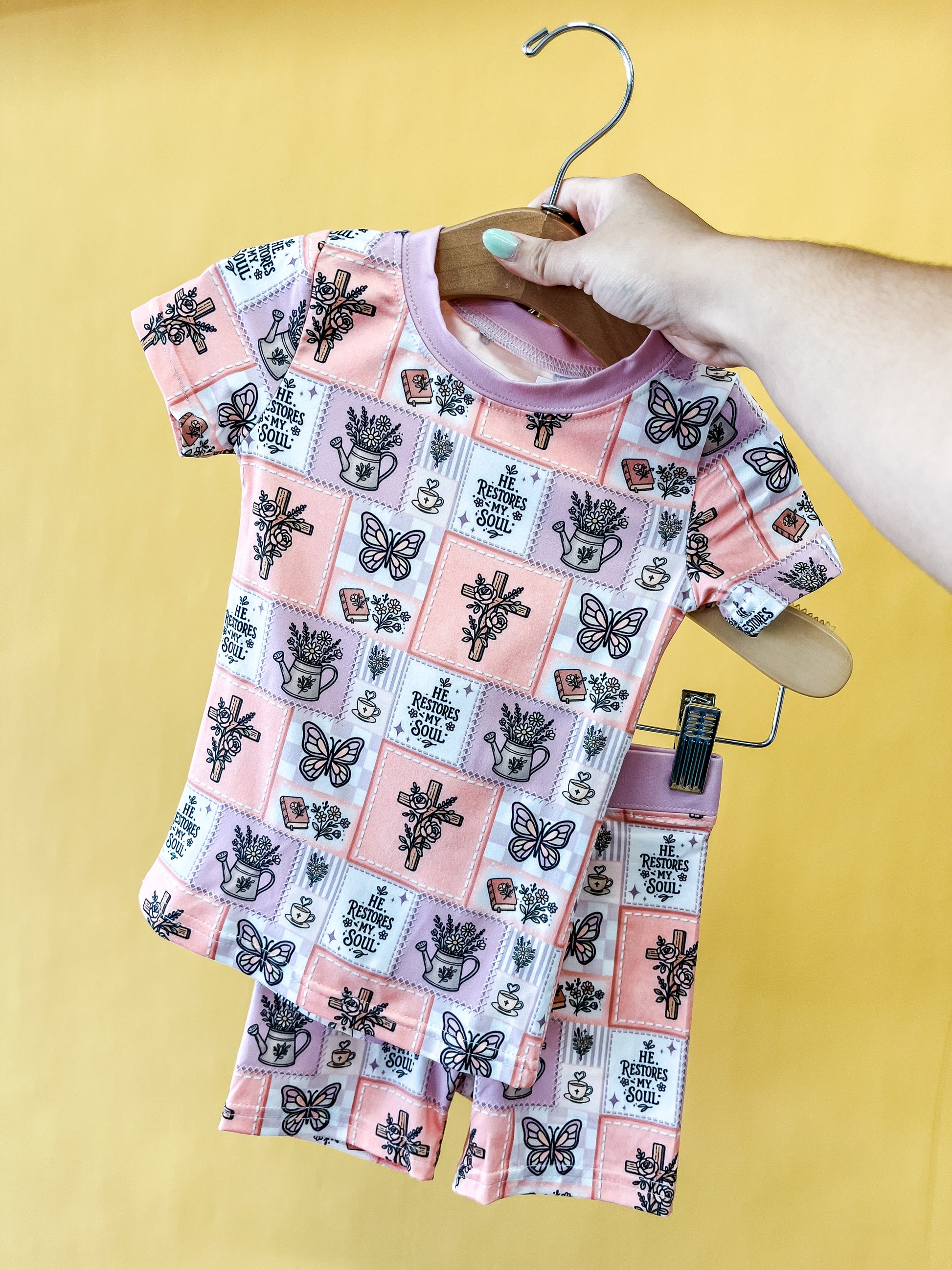 A hand holds a hanger displaying the Tiny Tot Bundles Faith In Bloom Lounge Set—a matching pink toddler shirt and shorts set with butterfly, flower, and “Free the Soul” prints—against a yellow background.