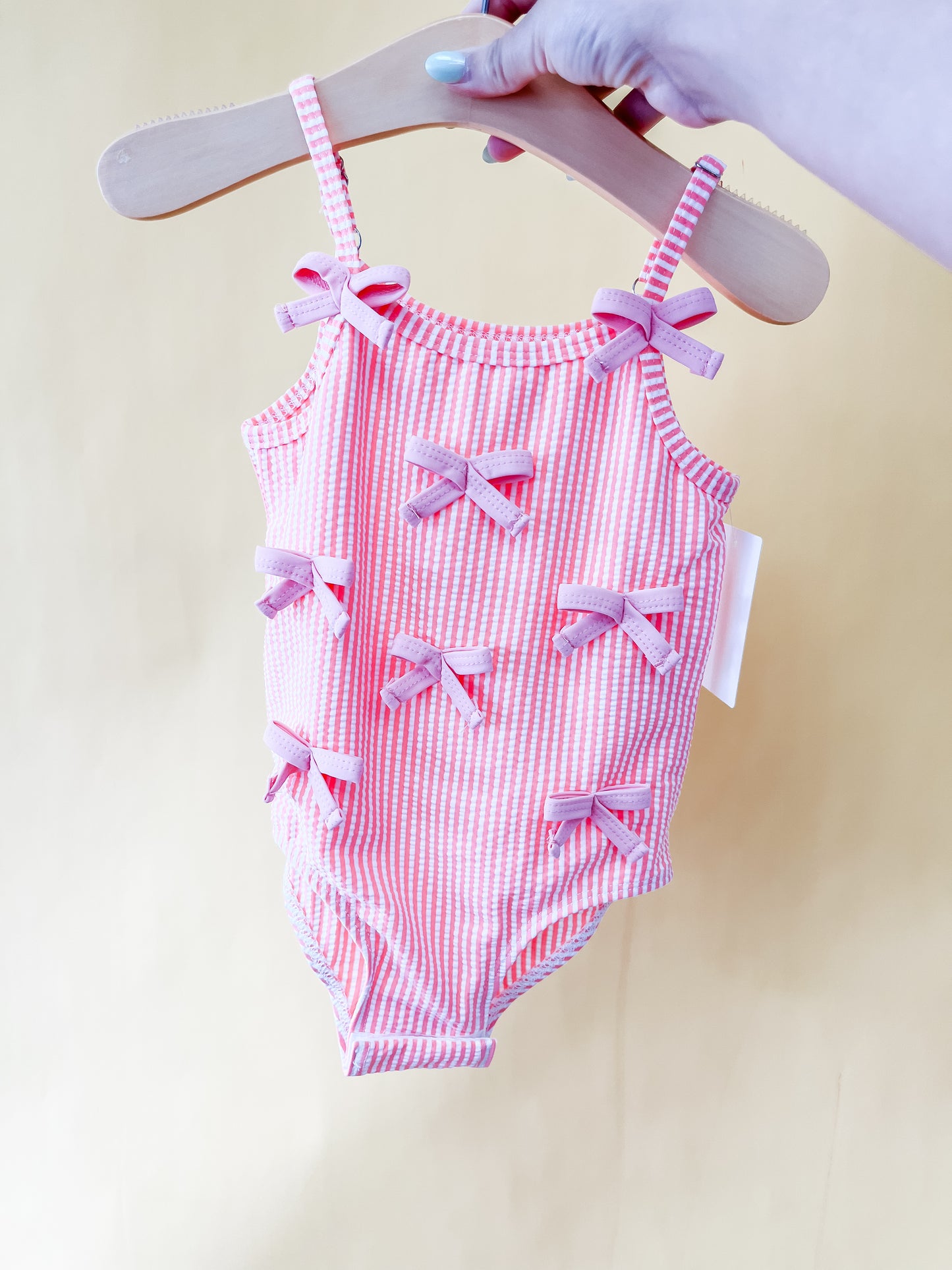 A hand holds the Pink Dainty Swimsuit by Tiny Tot Bundles—a pink and white striped toddler swimsuit adorned with six small pink bows—displayed on a wooden hanger against a plain light yellow background.
