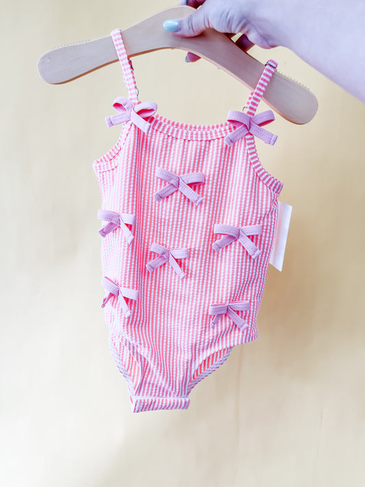 A hand holds the Pink Dainty Swimsuit by Tiny Tot Bundles—a pink and white striped toddler swimsuit adorned with six small pink bows—displayed on a wooden hanger against a plain light yellow background.