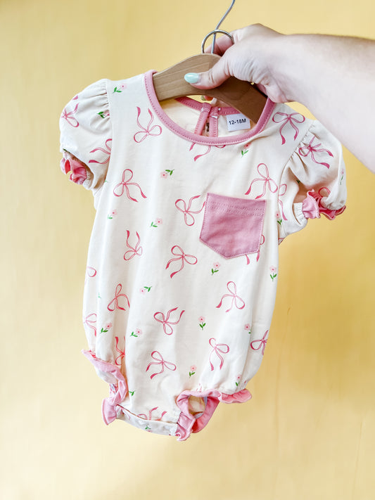 A hand holds the Tiny Tot Bundles Bow Dream Romper, a cream-colored baby outfit featuring pink trim, puffed sleeves, a pink chest pocket, and a charming pattern of pink bows and tiny flowers, displayed on a wooden hanger against a yellow background.
