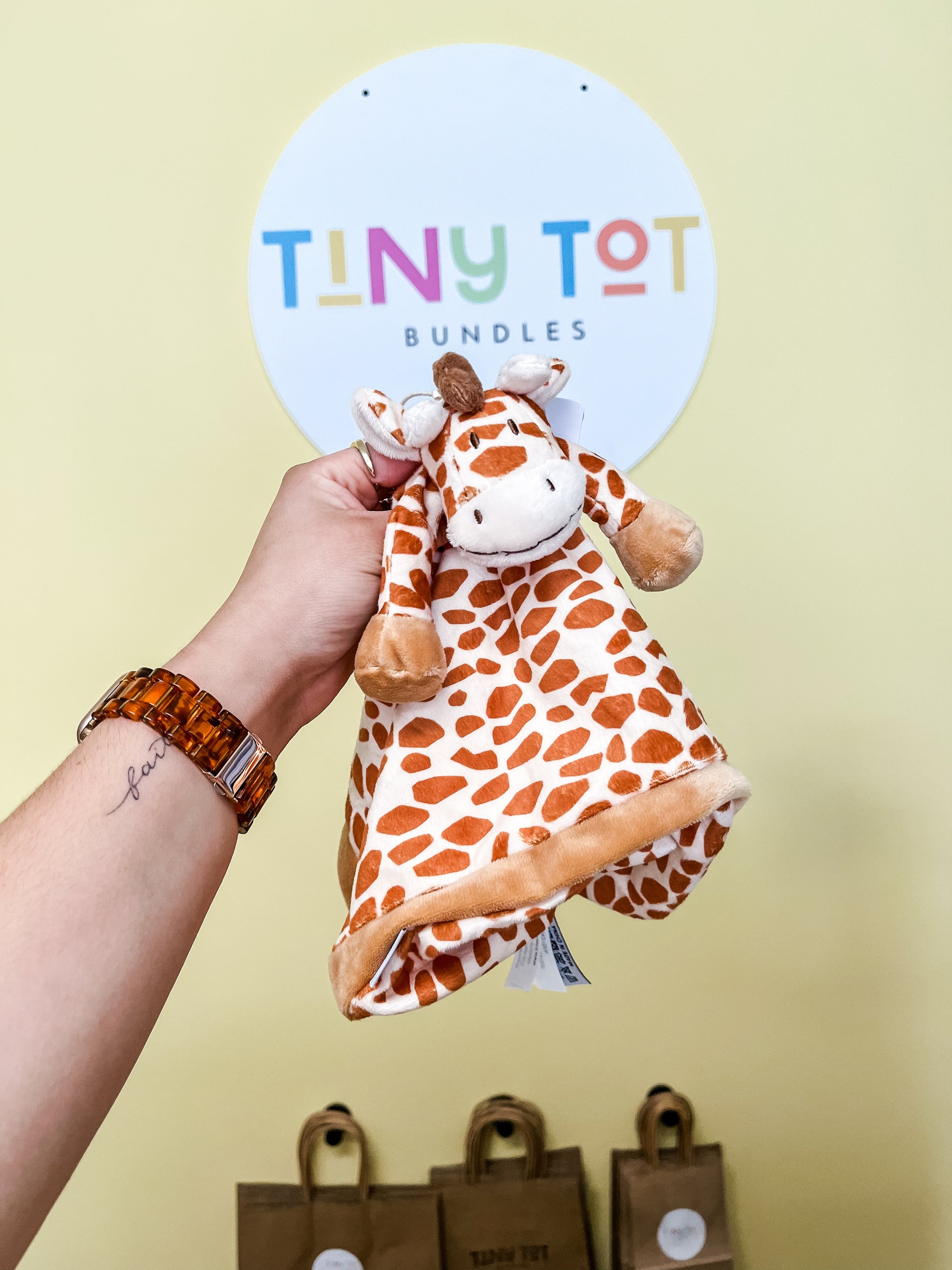 A hand holds a Snuggle Plush with a giraffe pattern, from Tiny Tot Bundles, in front of a sign with the brand name. Brown paper bags can be seen in the background against a yellow wall.