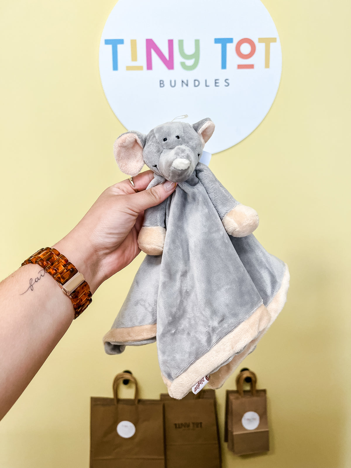 A hand holds a gray Snuggle Plush elephant blanket by Tiny Tot Bundles. Behind it, a round Tiny Tot Bundles sign hangs on a yellow wall beside brown paper bags with the same logo.