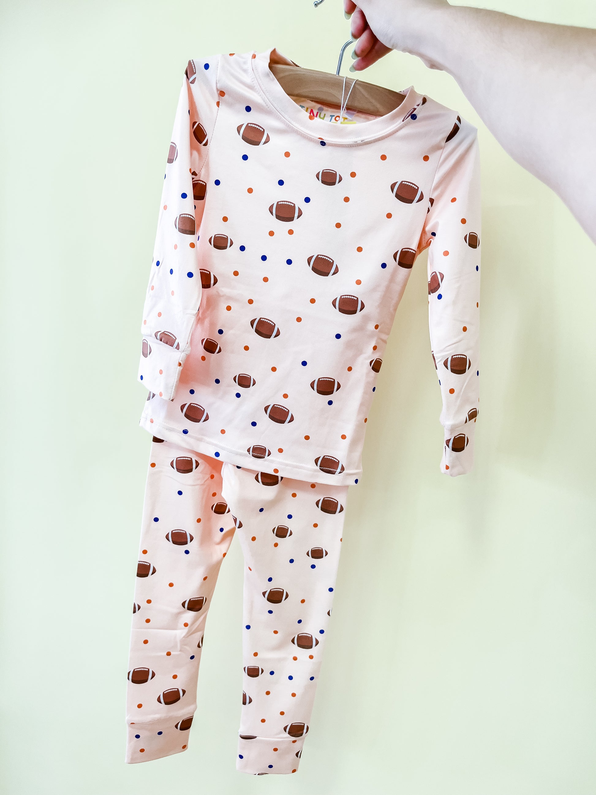 A hand holds up the Tiny Tot Bundles Football Frenzy Jammies Set on a hanger. The long-sleeve pajama set is light pink with a football and colorful dot pattern.