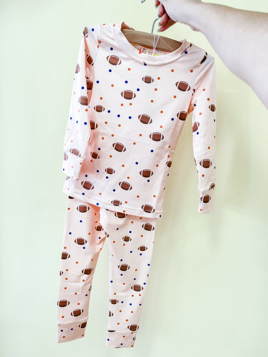 A hand holds up the Tiny Tot Bundles Football Frenzy Jammies Set on a hanger. The long-sleeve pajama set is light pink with a football and colorful dot pattern.
