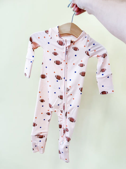 A hand holds a hanger with the Tiny Tot Bundles Football Frenzy Zippy, a baby onesie showcasing a pattern of brown footballs and blue and orange dots, displayed against a light background.