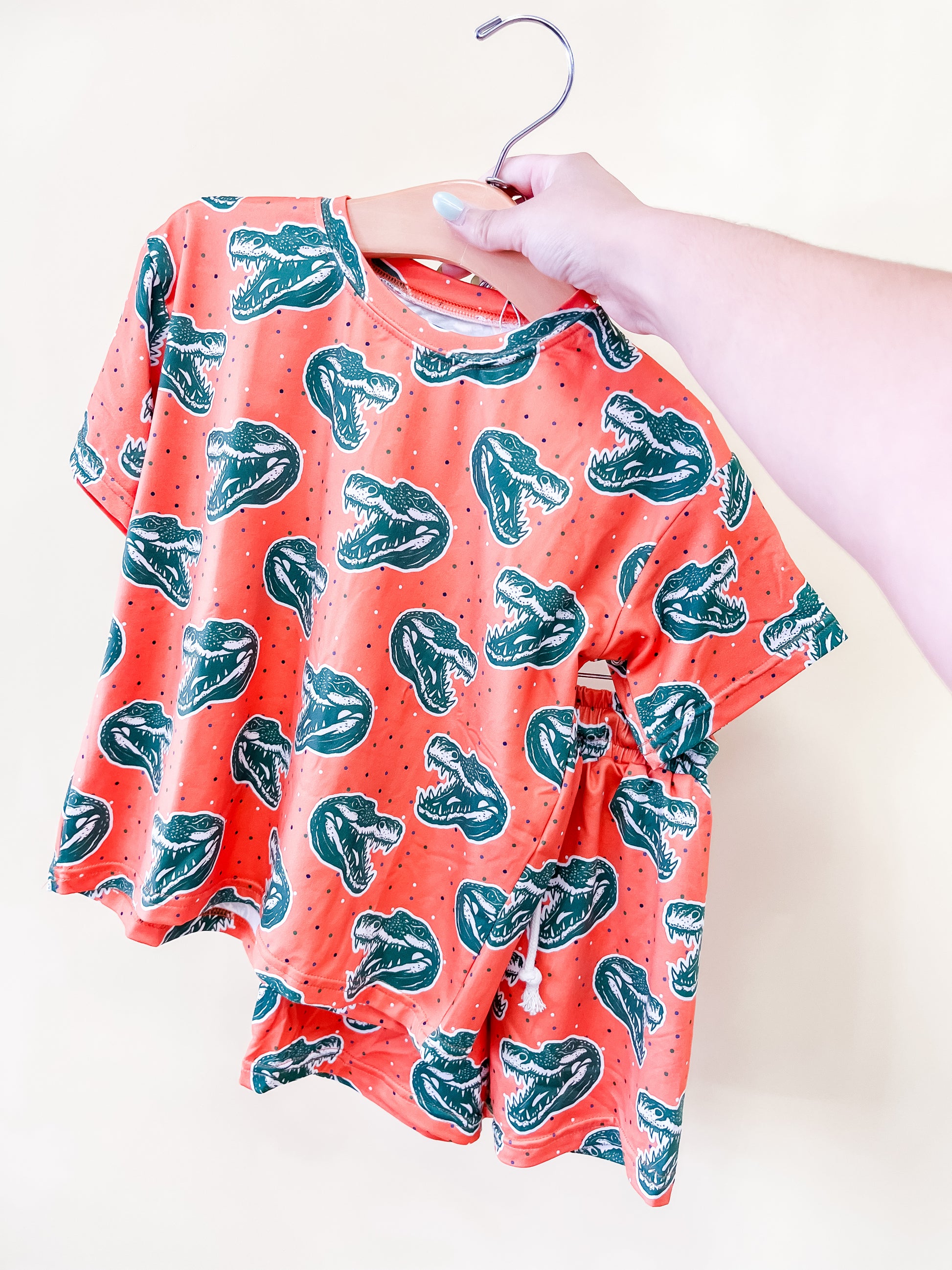 A hand holds up the Chomp Chomp Lounge Set by Tiny Tot Bundles, featuring a coral-colored T-shirt with a green dinosaur head print and small dots, displayed on a wooden hanger against a plain white background.
