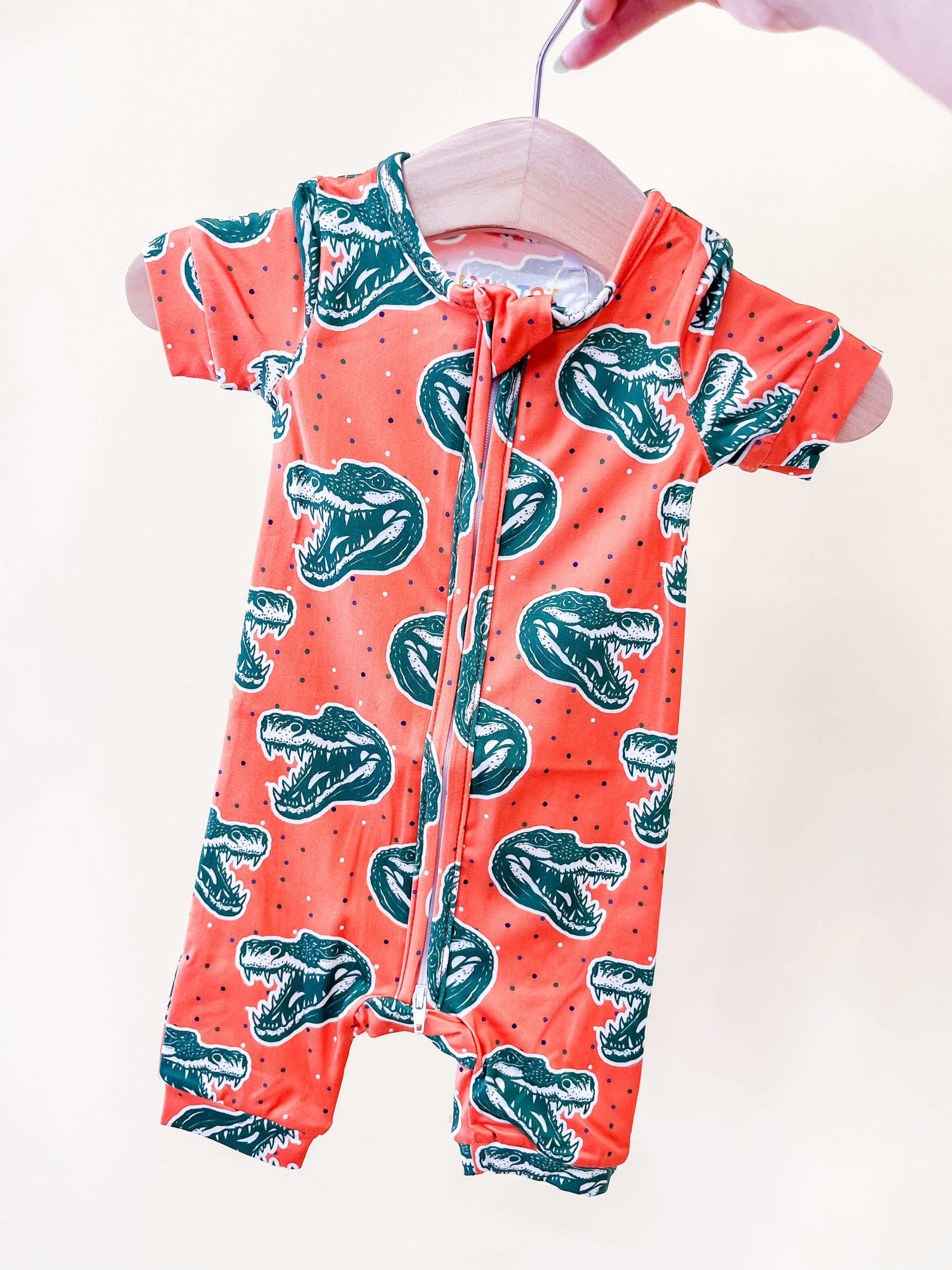A hand holds a wooden hanger displaying the Chomp Chomp Short Zippy by Tiny Tot Bundles—an orange baby romper adorned with green dinosaur head prints—set against a light background.
