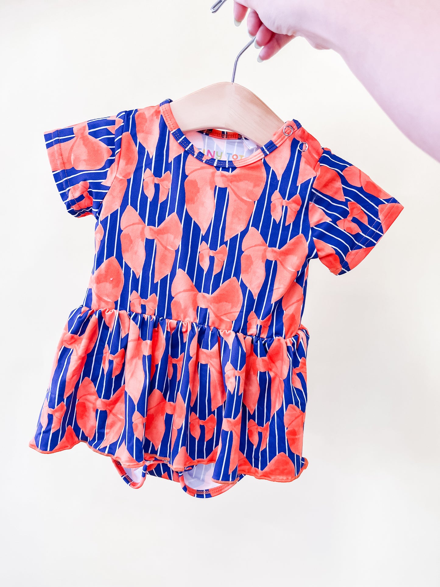 A hand holds the Tiny Tot Bundles Tailgate Bows Twirl Romper on a hanger. The romper is blue with vertical stripes and an orange floral pattern, featuring short sleeves and a ruffled skirt.