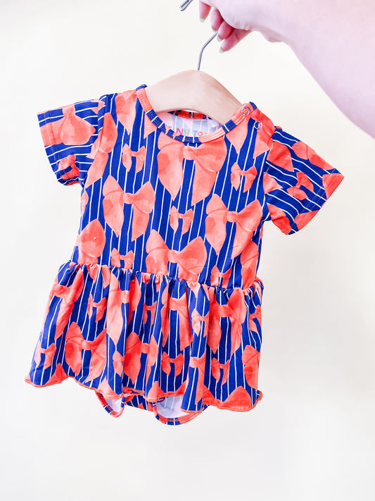 A hand holds the Tiny Tot Bundles Tailgate Bows Twirl Romper on a hanger. The romper is blue with vertical stripes and an orange floral pattern, featuring short sleeves and a ruffled skirt.