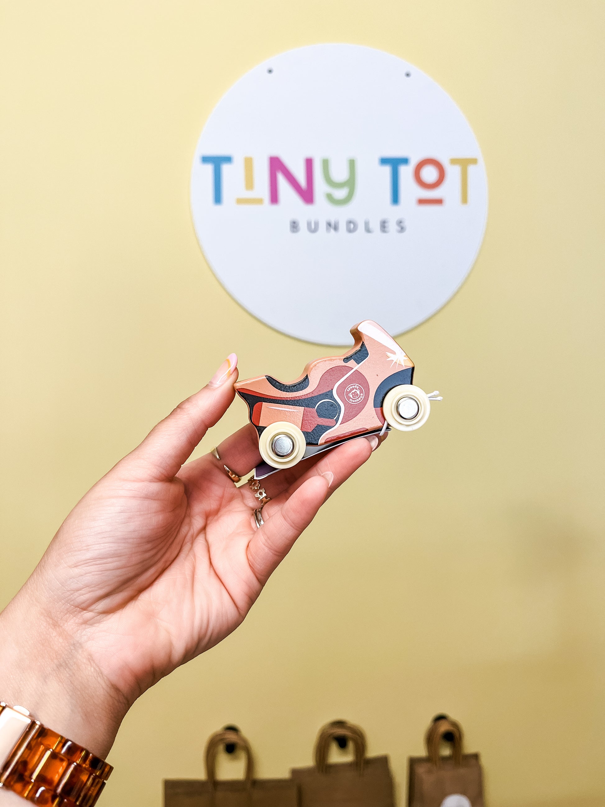 A hand holds a small Speedy Monkey Utility Vehicles Toy in front of a TINY TOT BUNDLES wall sign. Blurred brown paper bags are visible in the background.