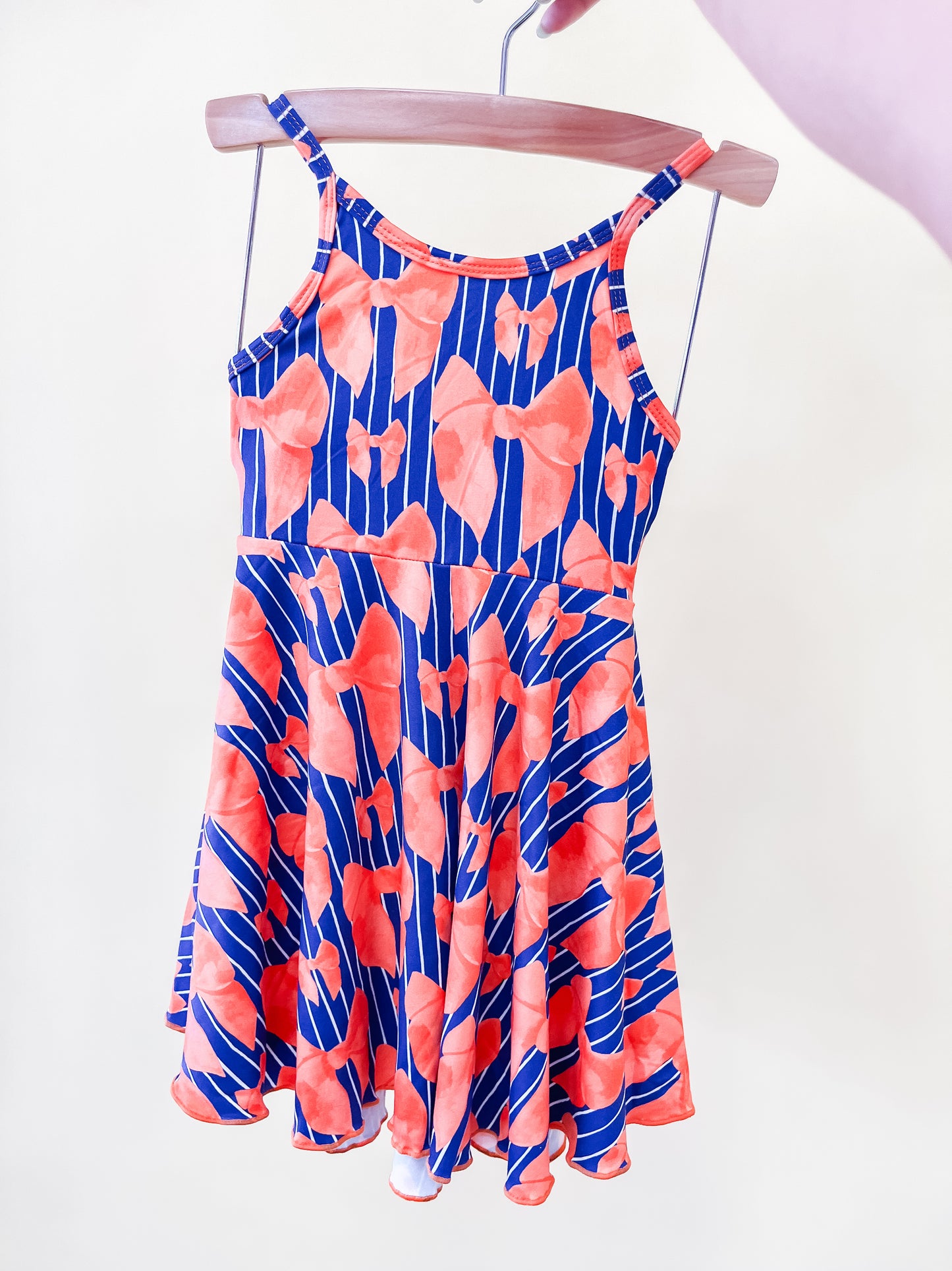 The Tailgate Bows Twirl Dress by Tiny Tot Bundles, a sleeveless dress with thin straps and a vibrant red and blue abstract pattern, is displayed on a wooden hanger against a light background.