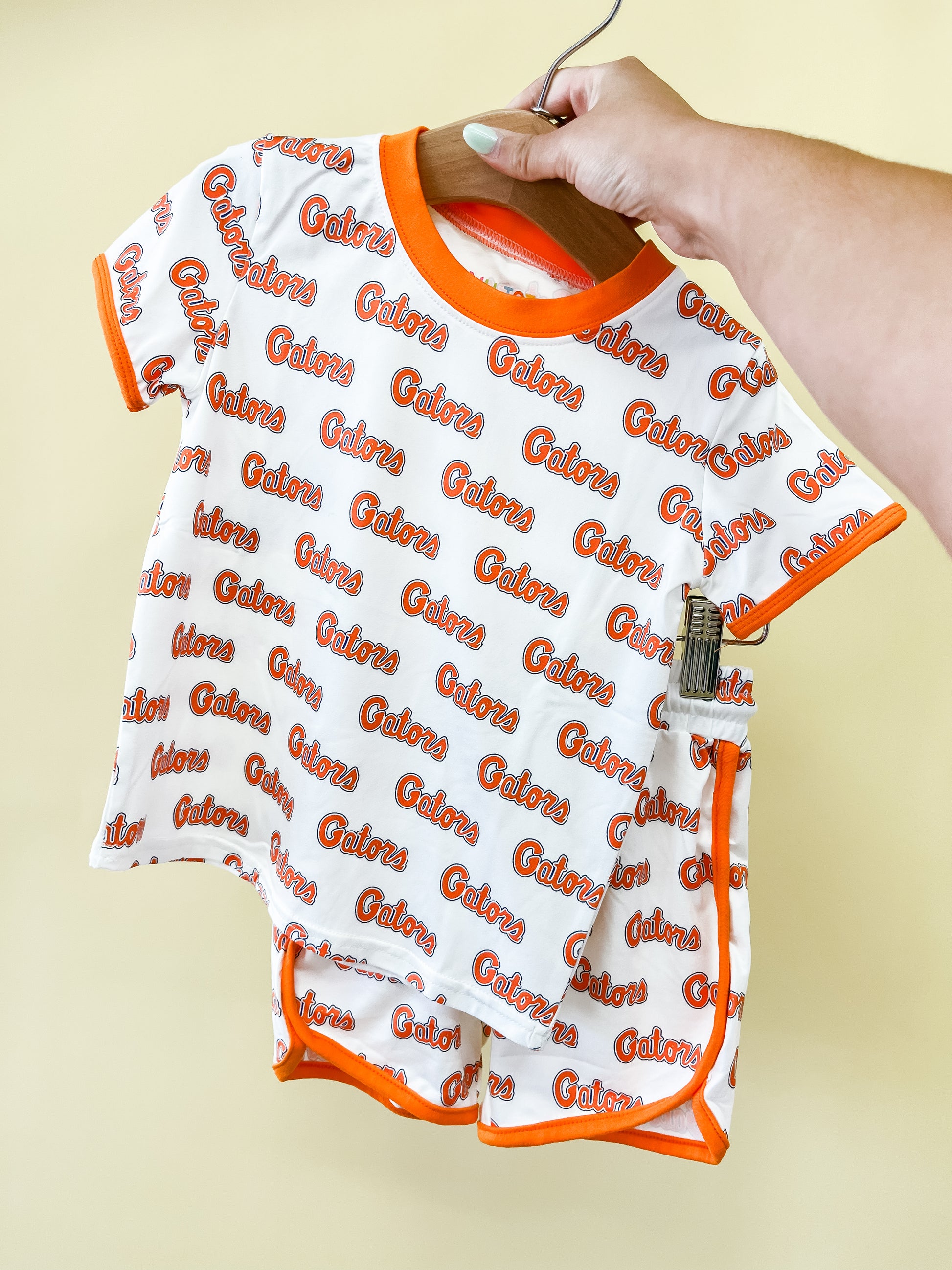 A hand holds up the Tiny Tot Bundles Go Gators Lounge Set, a matching children’s t-shirt and shorts set featuring orange trim and the phrase “Go Gators” printed repeatedly in orange and red on a white background.