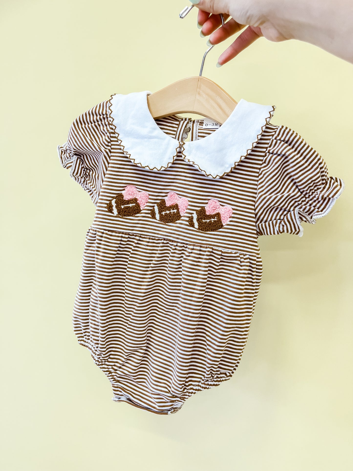 A hand holds the Friday Nights Bows Romper by Tiny Tot Bundles, featuring puff sleeves, a white scalloped collar, and charming embroidered bows with pink hearts across the chest, displayed on a hanger against a pale yellow background.
