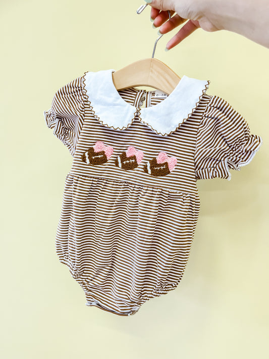 A hand holds the Friday Nights Bows Romper by Tiny Tot Bundles, featuring puff sleeves, a white scalloped collar, and charming embroidered bows with pink hearts across the chest, displayed on a hanger against a pale yellow background.