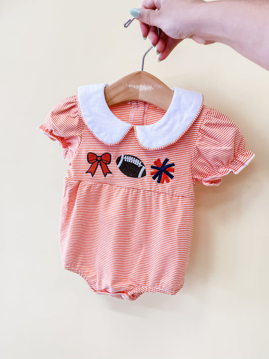 A hand holds a hanger with the Saturday Sweetheart Romper by Tiny Tot Bundles. The romper features orange and white stripes, a white collar, and embroidered designs of a bow, football, and cheer megaphone on the chest.
