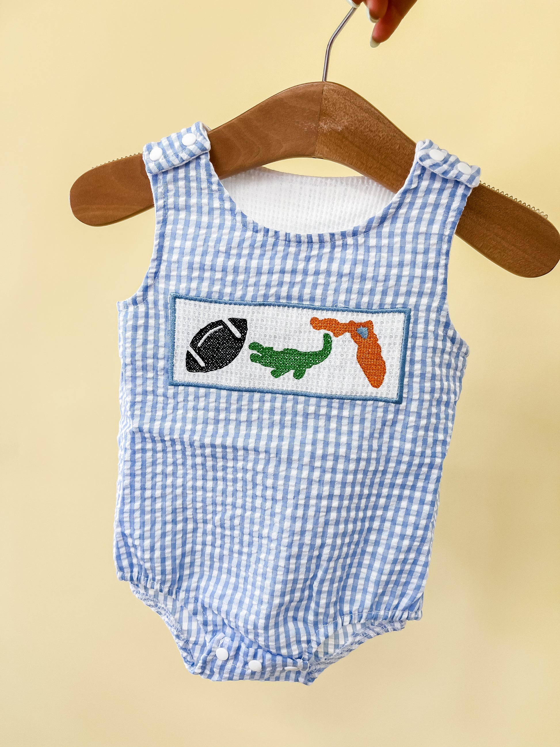 A blue and white gingham Sunshine State Tank Romper by Tiny Tot Bundles hangs on a hanger, featuring embroidered patches of a football, green alligator, and orange object on the chest.