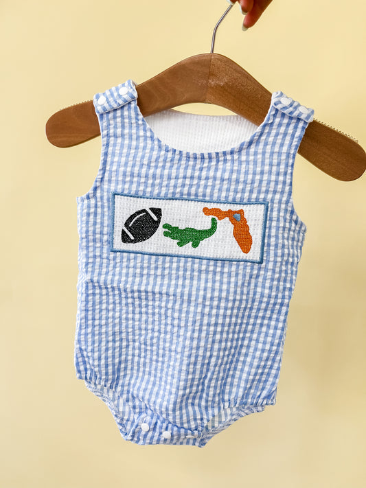A blue and white gingham Sunshine State Tank Romper by Tiny Tot Bundles hangs on a hanger, featuring embroidered patches of a football, green alligator, and orange object on the chest.