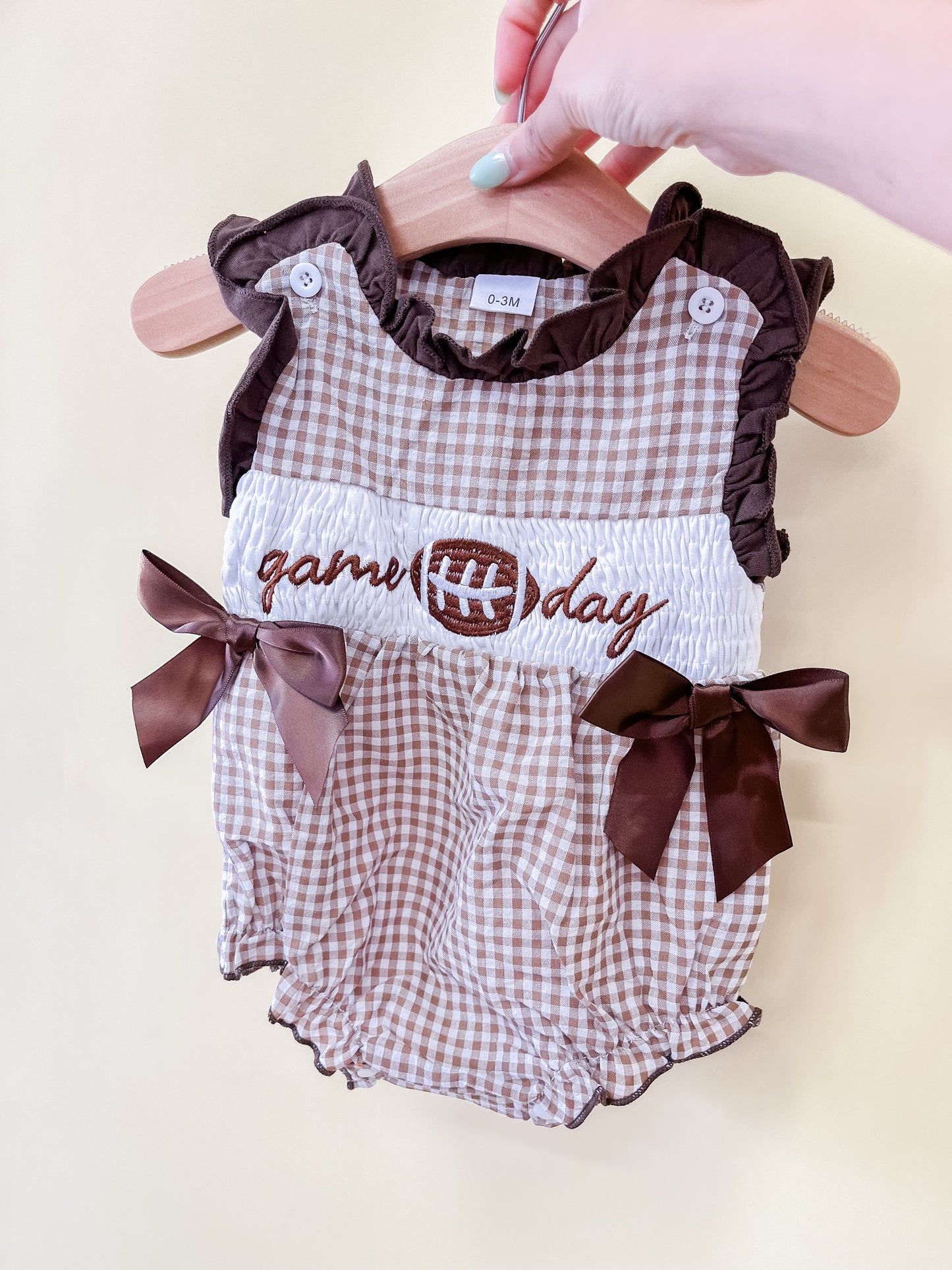 A hand holds a Tiny Tot Bundles Game Day Darlin Romper on a hanger. The brown and white checkered romper features ruffles, brown bows, and “game day” with a football embroidered on the front.