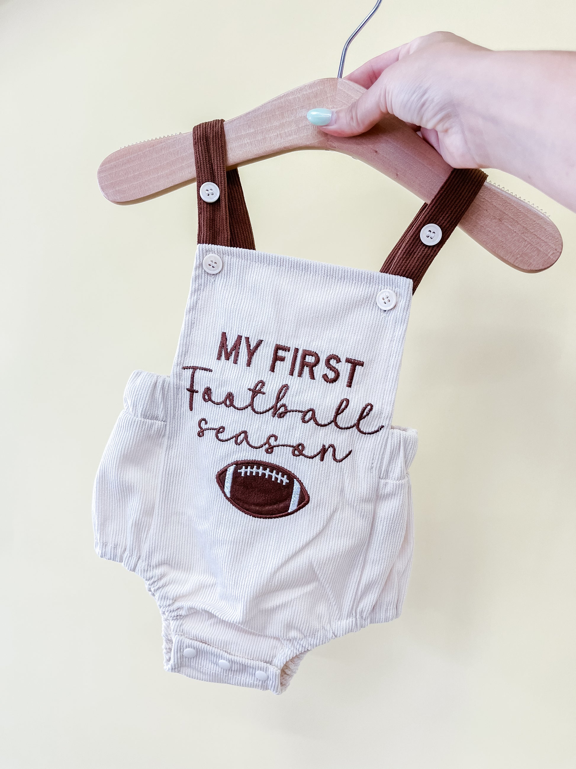 A hand holds a hanger with the Tiny Tot Bundles Rookie Season Overalls, featuring brown straps and “My First Football Season” with a football graphic on the front.