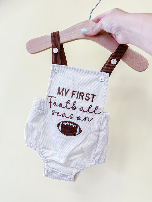 A hand holds a hanger with the Tiny Tot Bundles Rookie Season Overalls, featuring brown straps and “My First Football Season” with a football graphic on the front.