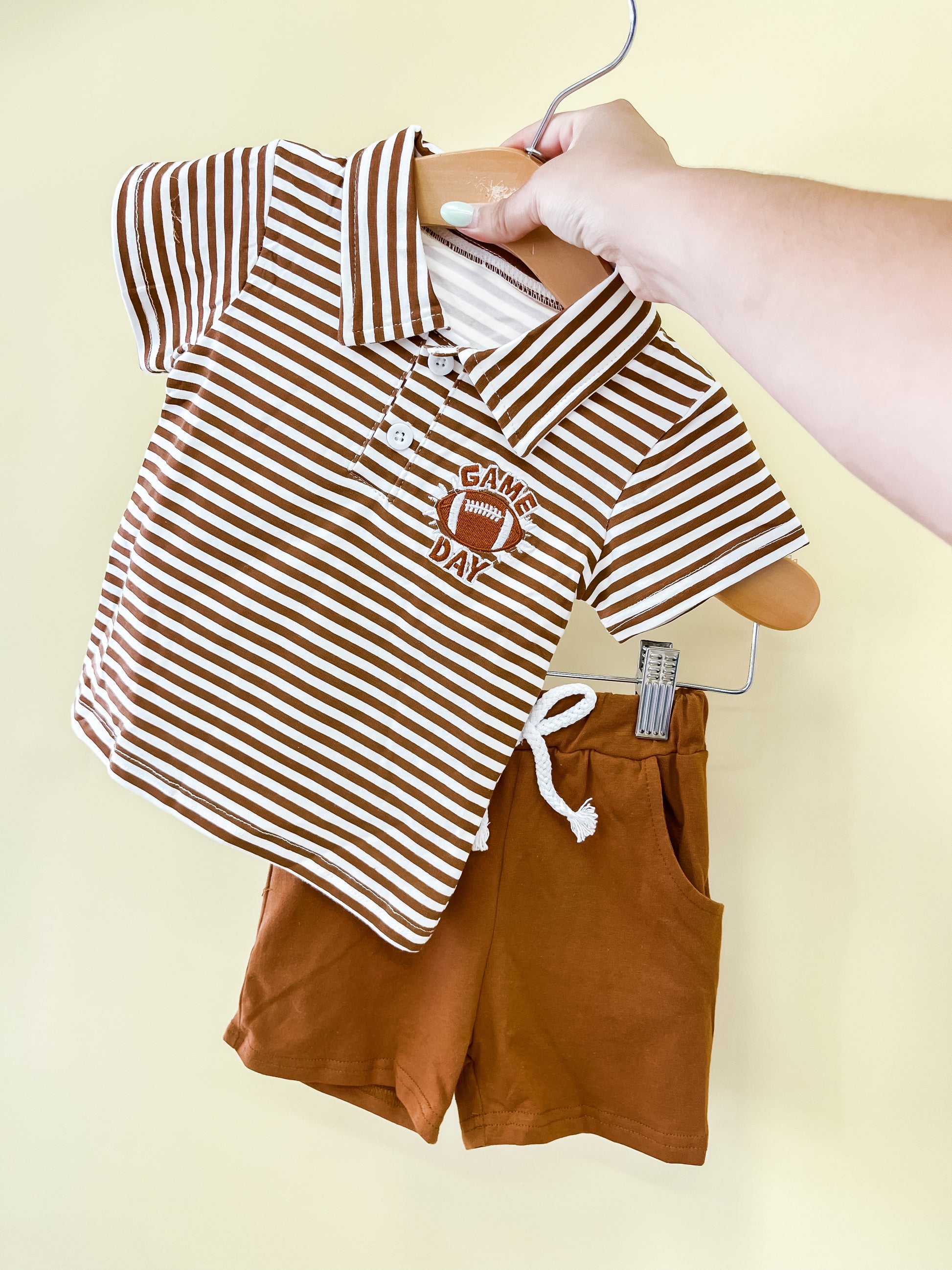 A hand holds the Tiny Tot Bundles Game Day MVP Short Set, featuring a brown and white striped polo with a football and “Game Day” applique, paired with matching brown shorts, displayed on wooden hangers against a light yellow background.