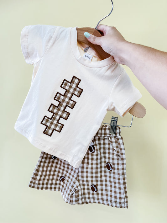 A hand holds up the Tiny Tot Bundles Kickoff Cutie Short Set, featuring a cream t-shirt with a football lace design and brown checkered shorts adorned with small football prints, against a pale yellow background.
