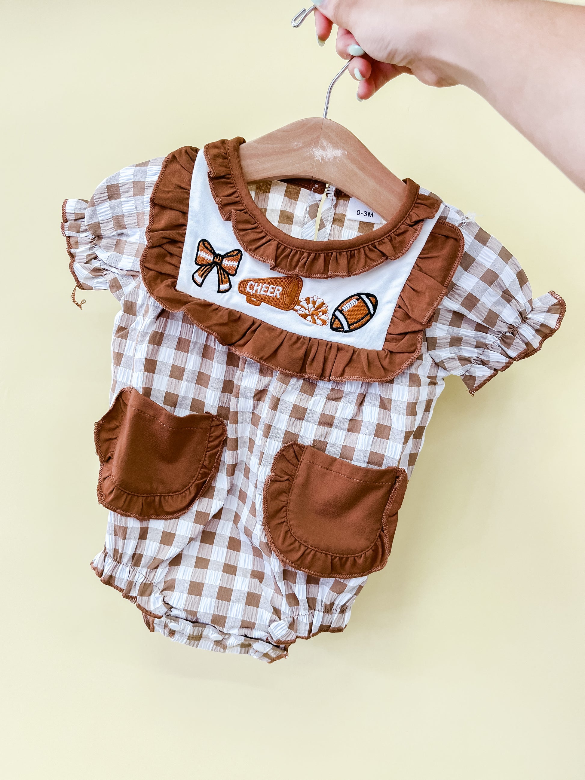 A hand holds the Tiny Tot Bundles Cheer Captain Romper, a brown and white checkered baby outfit featuring ruffled brown pockets and cheerleader-themed embroidered patches, including a megaphone and football, on the chest.