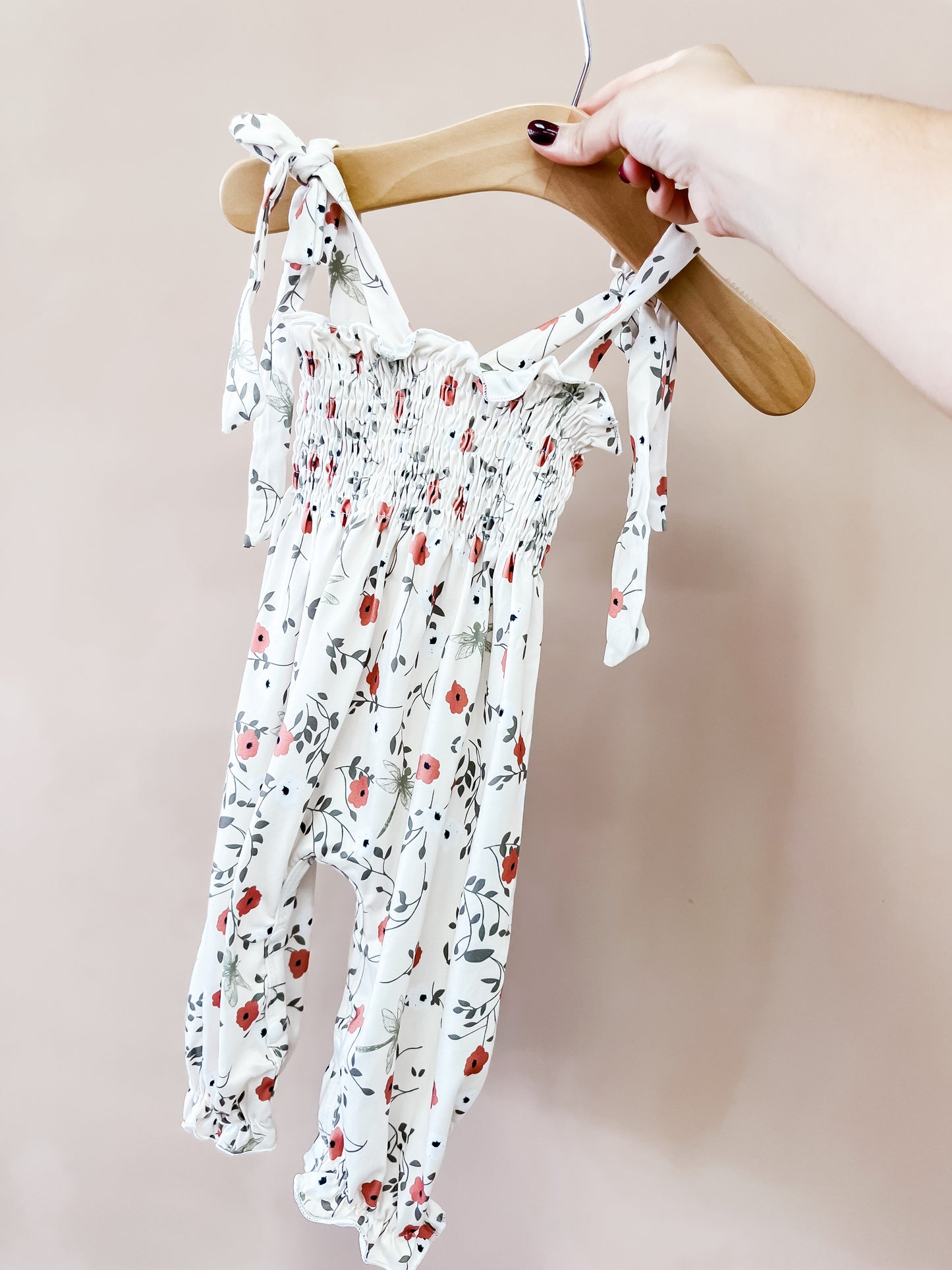 A hand holds a wooden hanger displaying the Tiny Tot Bundles “Rustic Blooms Jumpsuit,” a white baby romper adorned with a floral pattern and tied shoulder straps, set against a plain beige background.